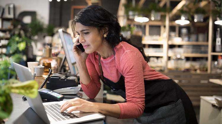 Woman who owns a small business start-up is on her cell phone in her flower shop typing on a laptop.